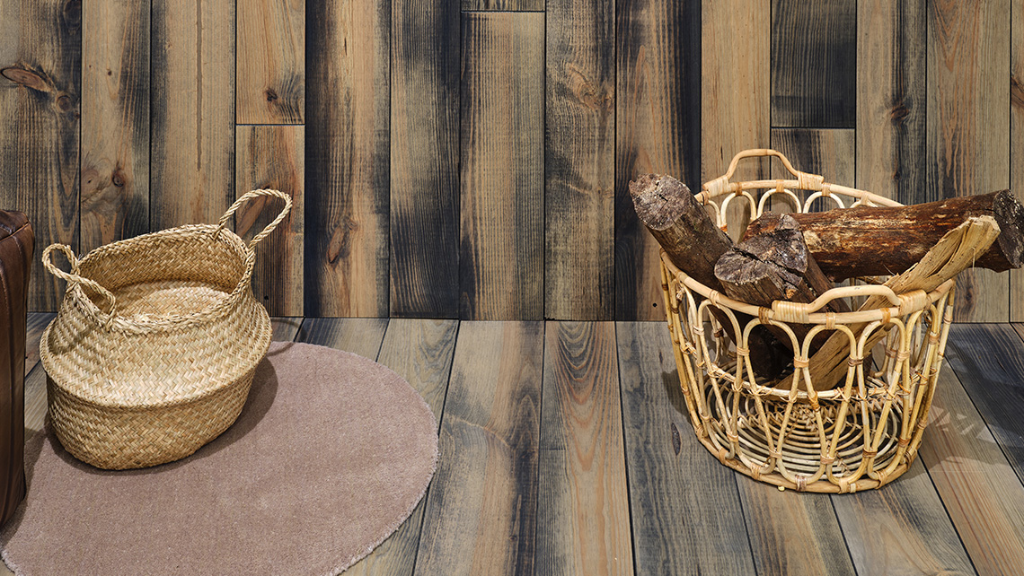 Un panier en osier rempli de bûches de bois posé sur un sol en bois. Un autre panier en osier vide est posé à côté sur un tapis rond. Le mur derrière est en bois foncé.